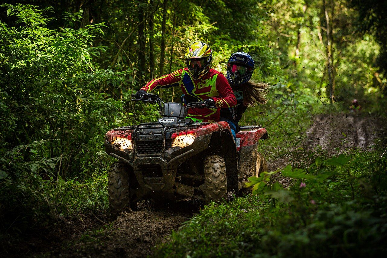 Illustrative photo taken in Brazil, not Bali. Two riders in helmets share one quad bike on a muddy jungle track